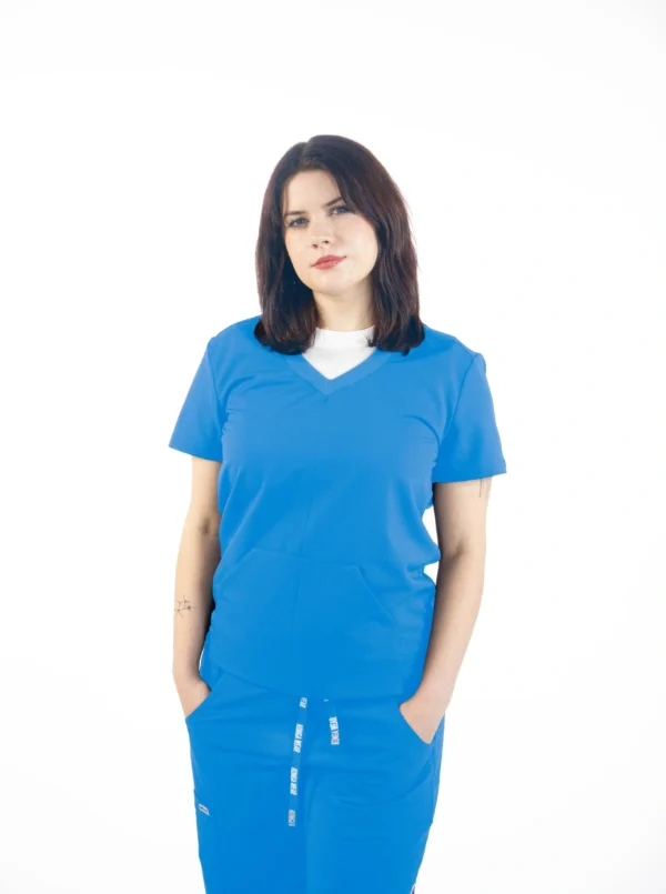 A woman in blue scrubs is standing with her hands in pockets.