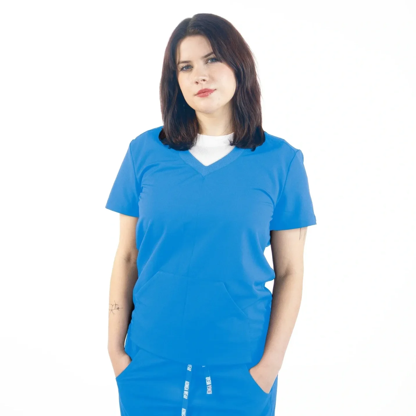 A woman in blue scrubs is standing with her hands in pockets.